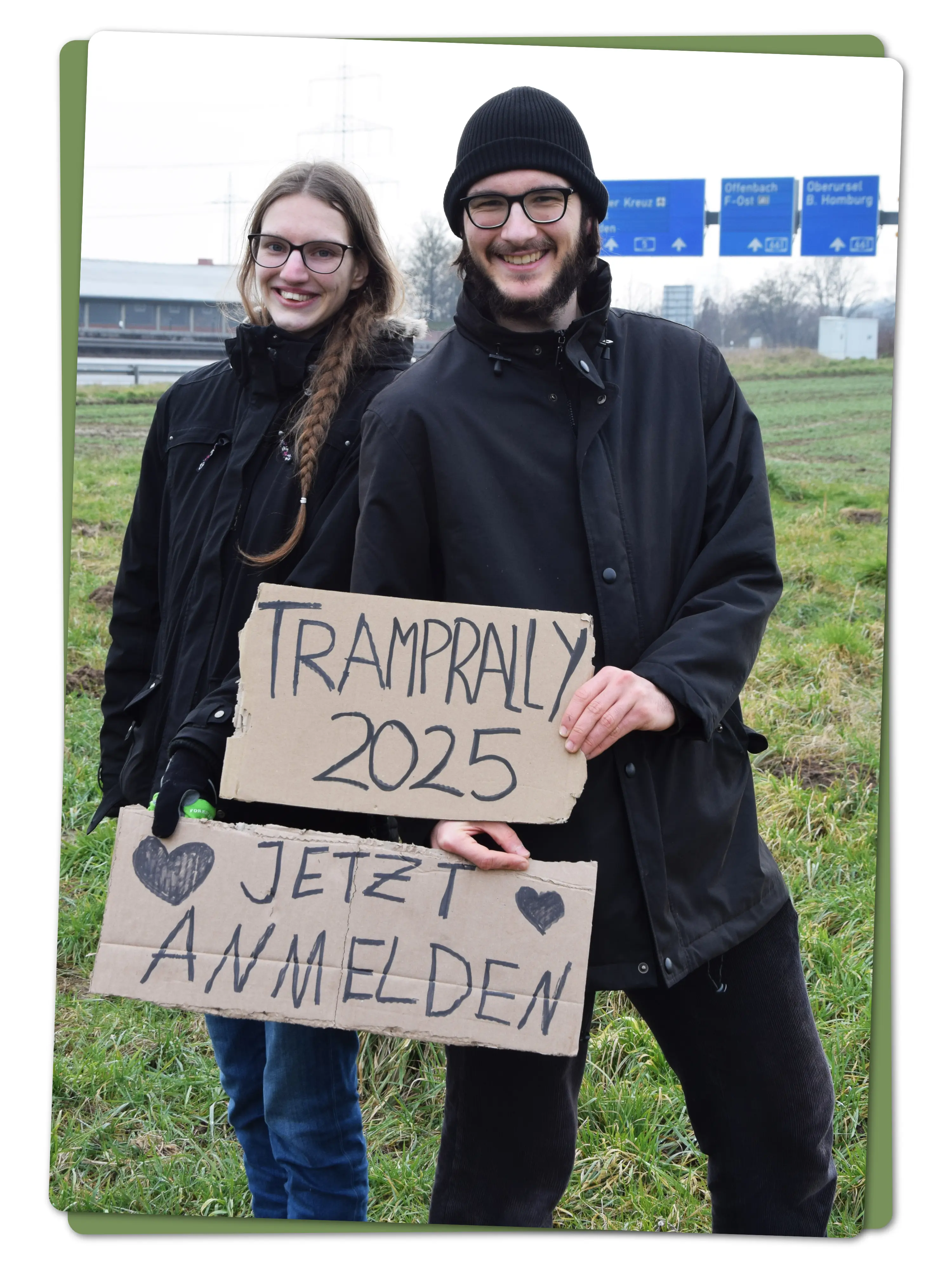 Zwei Tramper stehen in der Nähe einer Autobahn und halten handgeschriebene Pappschilder mit der Aufschrift "Tramprally 2025, Jetzt Anmelden"